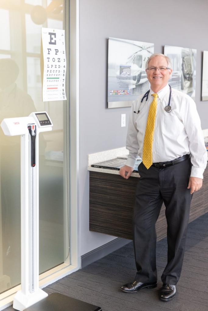 Dr. Steve Edmondson, DO, FAA AME (Aviation Medical Examiner) and Board-Certified in Family Medicine, standing next to his exam room near a scale and eye chart.